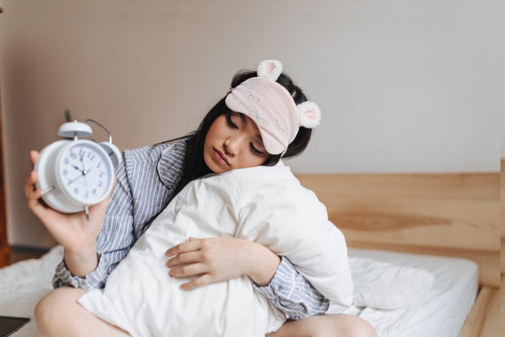girl in pink sleeping mask is hugging pillow and looking at alarm clock with sadness.