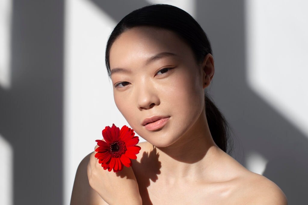Rutina K-beauty Mujer coreana con piel natural sosteniendo una flor roja con reflejo de la luz del sol y luminosidad en el rostro.